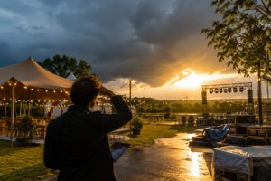 Organisateur observant le ciel avant un événement extérieur, avec une scène, des tentes et une météo changeante au coucher du soleil