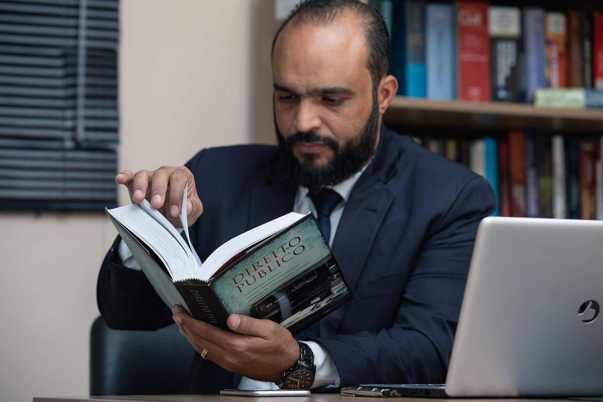 Homme en costume noir lit un livre, ordinateur portable à côté, étagères de livres en arrière-plan