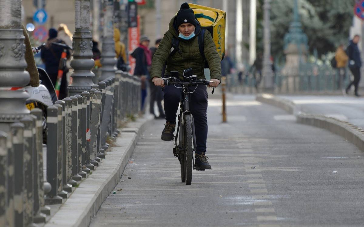 Livreur à vélo sur trottoir urbain, portant un colis, en manteau et casque